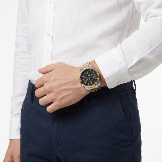 A close-up of a man wearing a two-tone gold and silver analog watch with a black dial, paired with a white linen shirt and navy blue trousers.