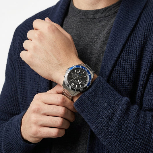 A close-up of a man wearing a silver stainless steel watch with a black dial and blue bezel, paired with a gray crewneck sweater and a navy blue knit cardigan.