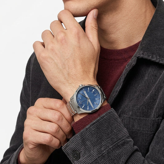 A close-up of a man wearing a silver-toned metal watch with a blue dial and white hour markers, styled over a burgundy sweater and dark gray corduroy jacket.