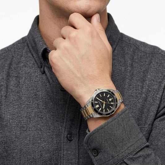 A close-up of a man wearing a two-tone stainless steel and gold watch with a black dial, paired with a dark gray textured button-down shirt.