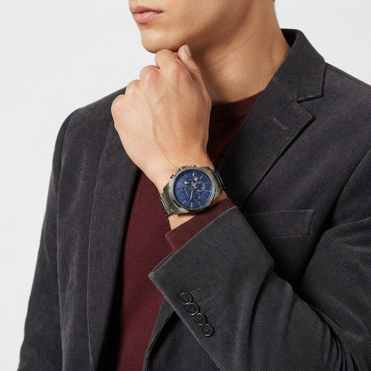 A close-up of a man wearing a silver-toned wristwatch with a blue dial and matching blue strap, styled with a dark gray blazer and burgundy sweater.