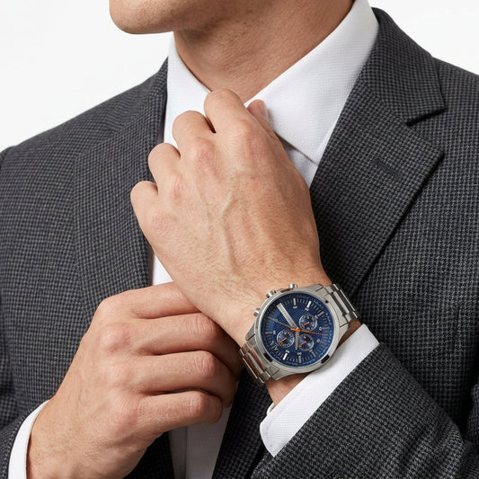A close-up of a man wearing a silver-toned metal watch with a blue dial and matching blue subdials, styled with a white dress shirt and a dark gray textured suit jacket.
