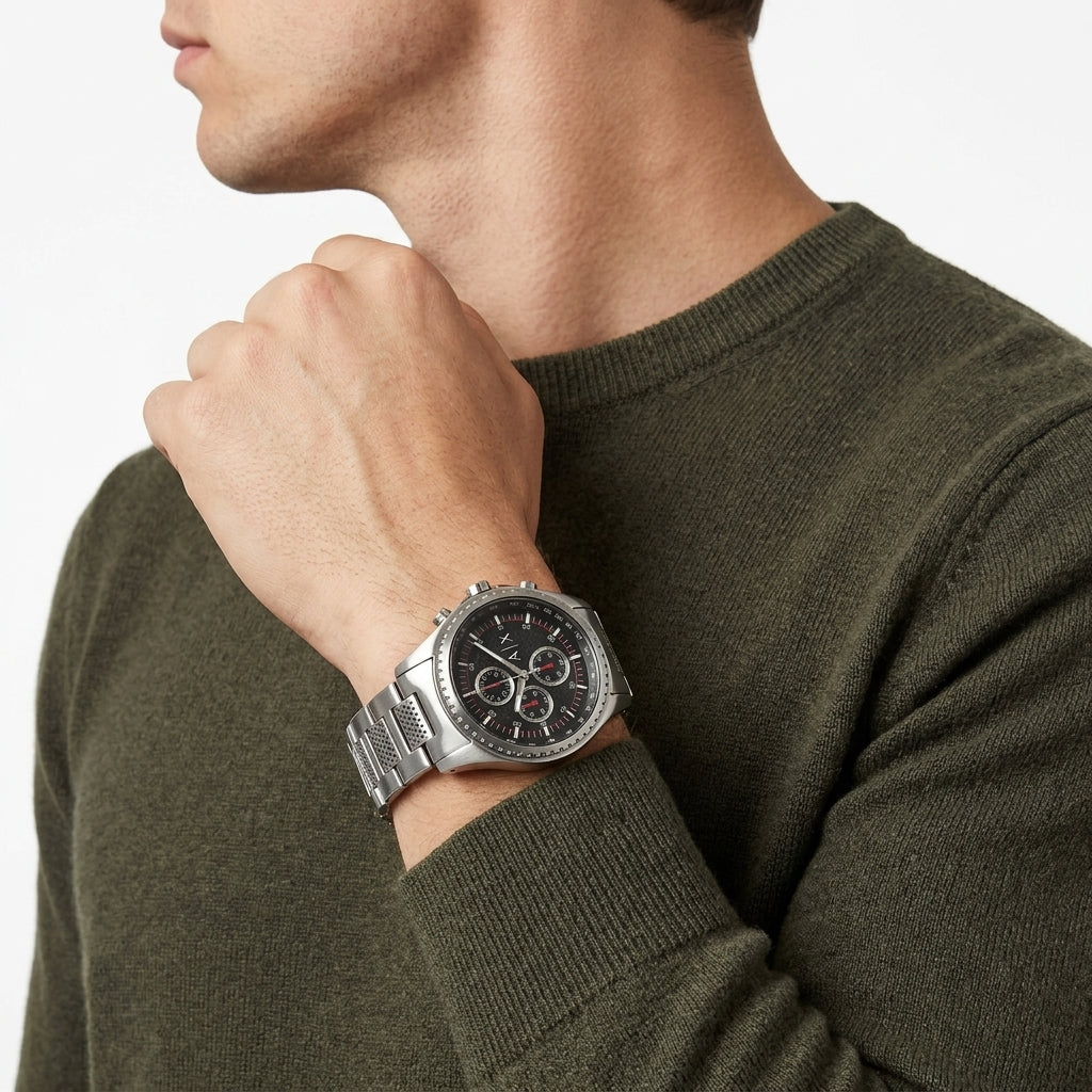 A silver stainless steel chronograph watch with a black dial and silver link bracelet, worn on a man's wrist. The man is wearing an olive green knit sweater against a clean white background.