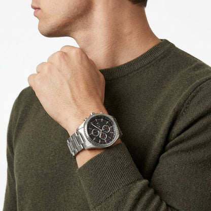 A silver stainless steel chronograph watch with a black dial and silver link bracelet, worn on a man's wrist. The man is wearing an olive green knit sweater against a clean white background.