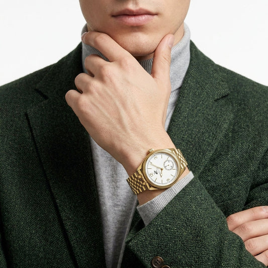 A close-up of a man wearing a gold-tone wristwatch with a white dial and matching gold bracelet, styled with a gray turtleneck and dark green textured blazer.