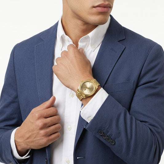 A man wearing a navy blue blazer and white dress shirt, wearing a gold-tone watch with a gold dial and matching gold-tone link bracelet.