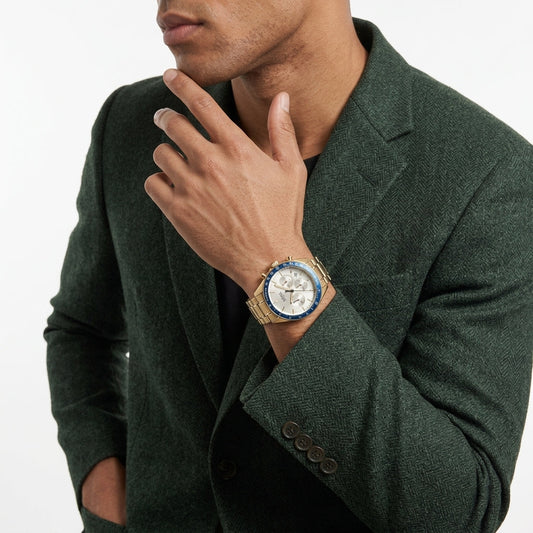 A man wearing a dark green textured blazer and a gold-tone wristwatch with a blue bezel and white dial, posed against a clean white background.