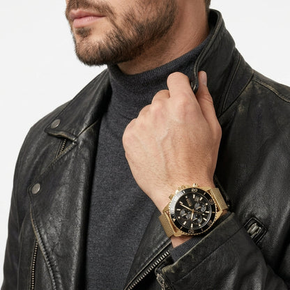 A close-up of a man wearing a gold-tone mesh watch with a black dial and bezel, styled with a black leather jacket and dark gray turtleneck sweater against a clean white background.