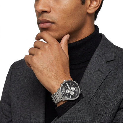 A close-up of a man wearing a silver stainless steel watch with a black dial and silver link bracelet, styled with a black turtleneck and gray blazer against a white background.