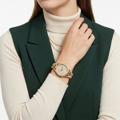 A close-up of a woman wearing a gold-tone watch with a round dial and matching metal link bracelet, styled over a cream turtleneck and dark green blazer vest.