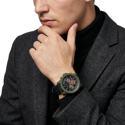 A close-up of a man wearing a dark gray blazer and black turtleneck, showcasing a rugged green analog watch with a black strap and orange accents on his wrist.