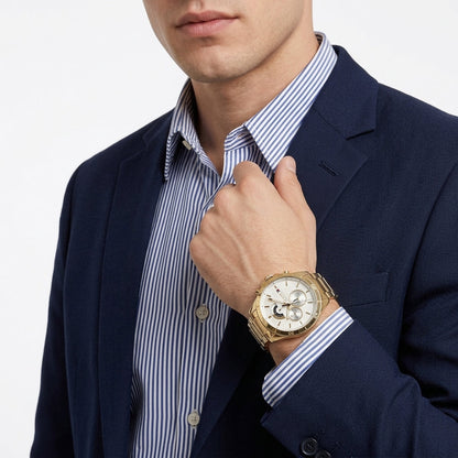 A man in a navy blazer and blue-and-white striped dress shirt wearing a gold-tone wristwatch with a white dial, shown in a close-up studio shot.