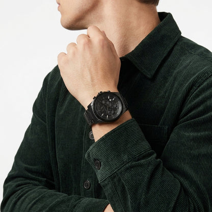 A man wearing a black multi-function wristwatch with a black strap, paired with a dark green corduroy shirt, shown in a close-up shot against a light background.