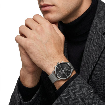 A close-up of a man wearing a silver mesh-strand watch with a black dial and silver case, styled with a black turtleneck and a gray herringbone blazer against a white background.