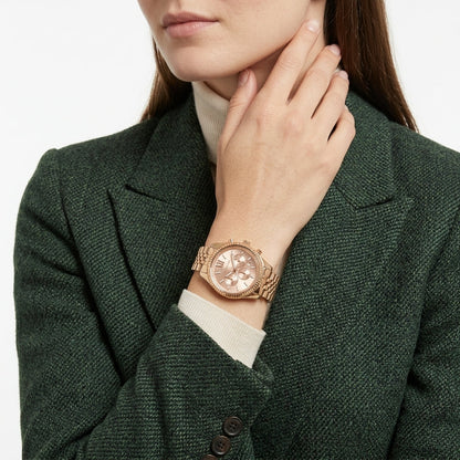 A close-up of a woman wearing a rose gold chronograph watch with a matching metal link bracelet, styled with a dark green textured blazer and a cream turtleneck.