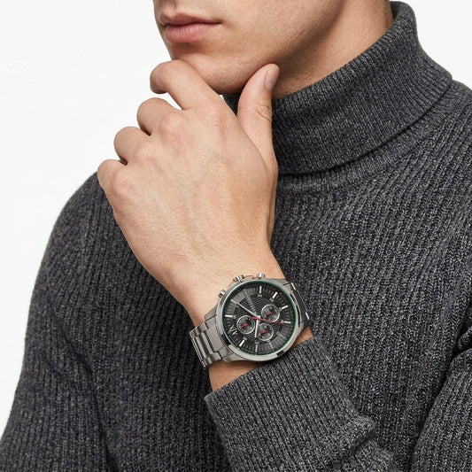 A close-up of a man wearing a silver-toned chronograph watch with a dark dial and metal link bracelet, paired with a dark gray ribbed turtleneck sweater.